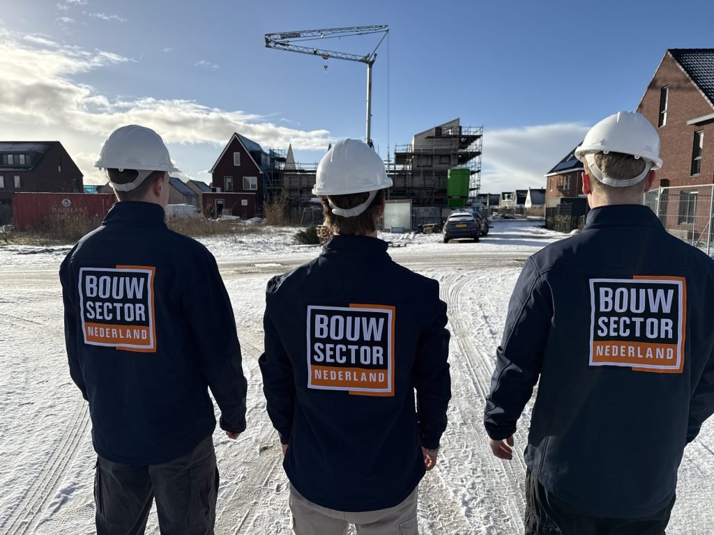 Team van Bouwsector Nederland in donkerblauwe jassen met oranje logo op de rug voor een bouwplaats